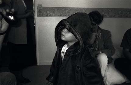  Enfant en manteau et tête recouverte d'une capuche avec femme en arrière-plan. Noir et blanc.