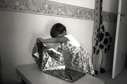  Paquet cadeau déballé par une petite fille en robe dans une chambre d'enfant. Noir et blanc.