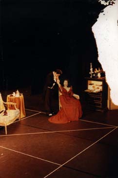  Plateau de théâtre avec scène de baise-main entre un homme en frac et une femme en robe longue rouge décolletée assise devant une coiffeuse. Image avec tache blanche.