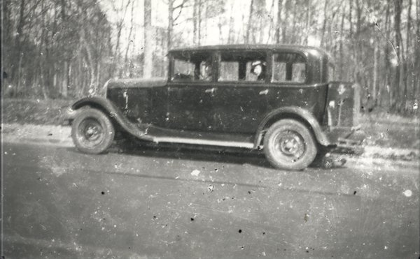 Citroën traction avant garée devant un bois avec une femme en chapeau à l’intérieur. Noir et blanc.