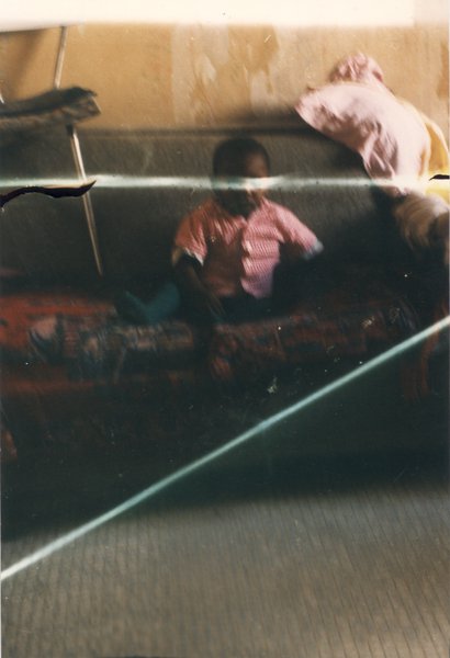Enfant de type africain assis sur un canapé où est posée une chaise. Image avec pliures.