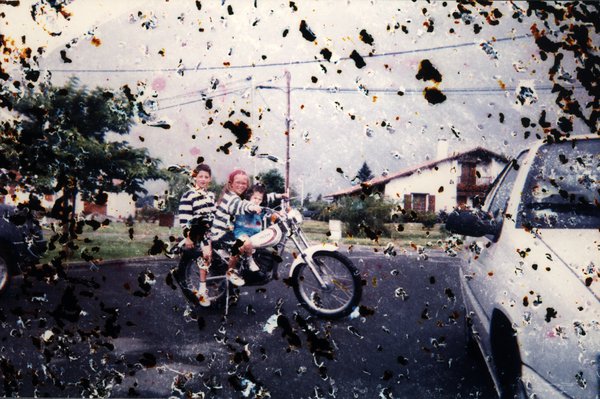 Trois enfants sur un vélomoteur dans une rue. Image très abîmée.ᴬᴵ
