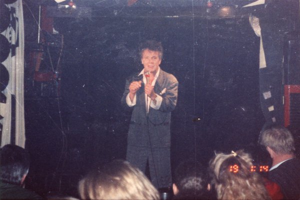 Chanteur en costume gris avec un micro sur une scène face à un public.