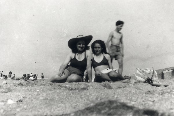 Deux jeunes filles assises en maillot sur une plage, jeune homme debout en arrière-plan. Noir et Blanc.